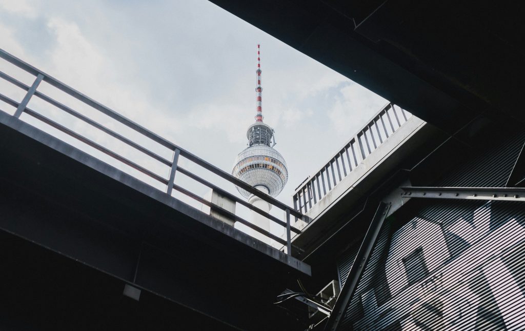 Kuppel des Berliner Fernsehturms, gerahmt durch zwei Abschnitte einer Brücke mit Geländern, Stahlträgern und Graffitis.